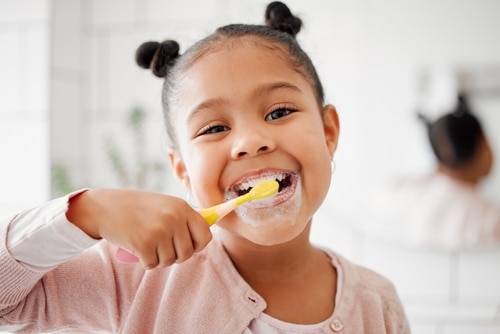 Young girl brushing her teeth with a yellow toothbrush, smiling, in a bright bathroom setting, emphasizing the importance of dental hygiene for children.